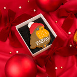 A decorated robin biscuit wearing a red winter bobble hat sitting in an open white Molly's Bakehouse gift box on red satin fabric with red bows, Christmas ornaments, and pearl-like beads around it. The iced biscuit has the word "name" written on it in white icing, suggesting personalisation.