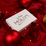 A white rectangular gift box with gold foil branding reading 'Molly's Bakehouse' lies on rich red satin fabric surrounded by red ornaments, bows, and small pearl-like beads. The product itself is not visible, so this is a packaging-focused shot.