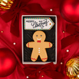 A decorated gingerbread man biscuit in a white gift box with a 'Merry Christmas' tag, surrounded by red satin bows, red ornaments, and gold holiday decorations.