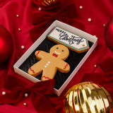 A gingerbread man Christmas biscuit in a white Molly's Bakehouse gift box with a black insert, placed on red satin background with Christmas holiday ornaments. The biscuit is decorated in beige, black and red icing with a red bow tie, red wavy trim, white buttons, and the text 'name' written across the chest waiting for personalisation