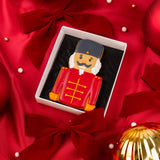 A nutcracker themed biscuit gift box in a with red velvet bows and ornaments on a red satin background.