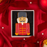 A nutcracker-shaped decorated cookie in a white gift box with red ribbon and ornaments around it.