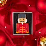 A hand decorated nutcracker themed iced biscuit in a white Molly's Bakehouse gift box, placed on red satin fabric with red bows and holiday ornaments.