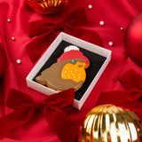 A bird-shaped Christmas cookie decorated with brown wings, a yellow-orange chest, and a red Santa hat with a white pom-pom, presented in a white gift box on a red satin background with bows and holiday ornaments.