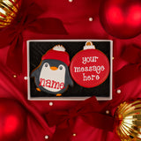A boxed holiday cookie set on red satin background with Christmas ornaments, featuring an iced penguin cookie in a red hat next to a bauble iced biscuit with the text 'your message here' ready to be personalised.