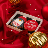 Two decorated Christmas cookies in a white gift box on red satin fabric with red bows and gold ornaments. One cookie is shaped like a penguin wearing a red Santa hat with the name 'name' written on it.