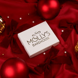 A white rectangular gift box printed with the brand name 'MOLLY'S BAKEHOUSE' in black, styled on a rich red satin background with red ribbons, pearl beads, and shiny red and gold Christmas ornaments.