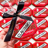 A hand holds a packaged Claridges London cookie in clear cellophane with a black ribbon, with multiple similar cookies arranged in the background.