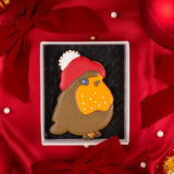 A decorated Christmas robin iced biscuit in a white Molly's Bakehouse gift box with a food grade cushion pad, placed on red satin fabric with bows and holiday ornaments.