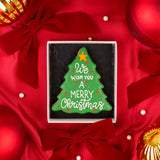 A Christmas tree-shaped hand iced biscuit with white royal icing text reading 'We Wish You A Merry Christmas' and small red iced decorative dots, presented in a luxury Molly's Bakehouse gift box on a red satin background with bows and Christmas holiday ornaments.