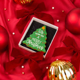 A Christmas tree-shaped iced biscuit gift with 'We wish you a Merry Christmas' written on it, presented in an open white gift box on a red background, surrounded by red velvet bows, red ornaments, and gold decorative elements.