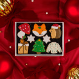 A boxed set of Christmas-themed iced cookies arranged on a molly's Bakehosue gift box, surrounded by red satin fabric, bows, Christmas ornaments, and edible earl-like beads. The Christmas biscuits include a red mushroom, fox face, hedgehog in a Santa hat, snowflakes, Christmas tree, and sweater and a Christmas present.