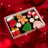 A white rectangular gift box filled with decorated Christmas-shaped sugar cookies, including a gingerbread man, candy cane, stocking, Christmas tree, gift box, and sweater cookie, set on red satin fabric with bows and pearl-like beads.