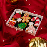 A white rectangular box filled with decorated Christmas iced sugar cookies, including a gingerbread man, stocking, candy cane, tree, gift box, and sweater cookie, arranged on a red satin fabric with ribbon bows and gold ornaments.