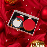 A white Molly's Bakehouse gift box containing two Christmas-themed cookies: a penguin cookie wearing a red hat and a bauble cookie, displayed on red satin fabric with bows and holiday ornaments.