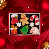 A top-down view of a Christmas-themed cookie gift box containing 8 decorated gingerbread cookies shaped like a stocking, gingerbread man, candy cane, gift box, Christmas tree, and Christmas sweater, set against a red satin background with holiday ornaments.