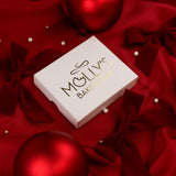 A white luxury Molly's Bakehouse gift box with gold foil branding sitting on a red satin fabric decorated with red ribbon bows, pearl-like beads, and glossy red ornaments.