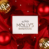 A white rectangular box with the logo 'MOLLY'S BAKEHOUSE' centered on a rich red satin background decorated with red and gold Christmas ornaments and dark red bows.