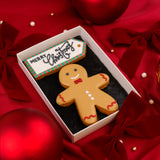 A gingerbread man Christmas biscuit in a white Molly's Bakehouse gift box with a black insert, placed on red satin background with Christmas holiday ornaments. The biscuit is decorated in beige, black and red icing with a red bow tie, red wavy trim, white buttons, and the text 'name' written across the chest waiting for personalisation