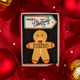 A gingerbread man Christmas biscuit in a white Molly's Bakehouse gift box with a black insert, placed on red satin background with Christmas holiday ornaments. The biscuit is decorated in beige, black and red icing with a red bow tie, red wavy trim, white buttons, and the text 'name' written across the chest waiting for personalisation