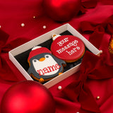Two decorated Christmas iced biscuits in a white Molly's Bakehouse gift box on a red satin background. One biscuit is a personalised penguin wearing a red winter hat with the text 'name' in red icing and the second biscuit is a personalised bauble with the text 'your message here' hand iced on it.