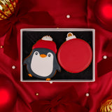 A boxed Christmas holiday biscuit set on a red satin background. The set includes a penguin biscuit wearing a cute red hat and a plain red bauble cookie both ready to be personalised. Both displayed in a white Molly's Bakehouse gift box with a black food grade cushion pad.